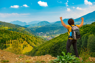 Fototapeta na wymiar Young mountain hiker with arms raised up, celelbrating and enjoying a beautiful mountain landscape covered with lush forests. Hiking in a sunny summer day.