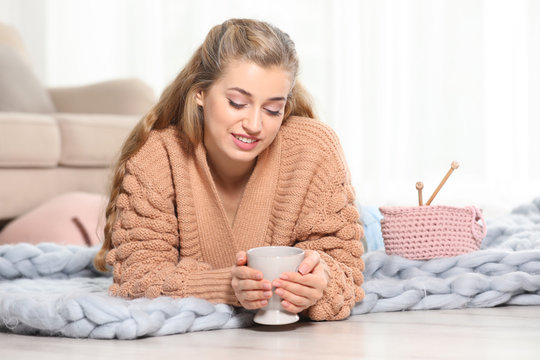 Attractive smiling young woman in cozy warm sweater with cup of hot drink lying on floor at home. Space for text