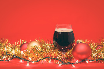 Dark ale beer in a tulip glass with christmas baubles tinsel and lights on red background