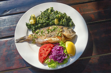 Balkan cuisine. Grilled fish  ( sea bream ) with vegetables  and slice of lemon on white plate. Dark rustic background, flat lay
