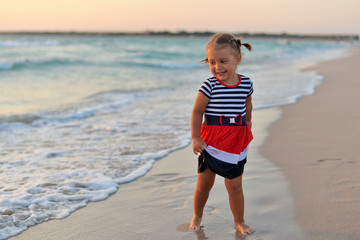 Happy little girl standing barefoot on the wet sand on the beach at sunset.