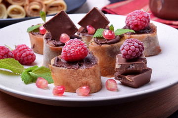 Rolled crepes with chocolate paste topped with raspberries, pomegranate, mint and chocolate pieces on a white plate