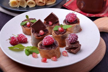 Rolled crepes with chocolate paste topped with raspberries, pomegranate, mint and chocolate pieces on a white plate