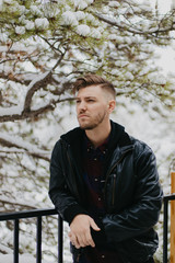 Portrait of Attractive Trendy Modern Young Male Model Smiling and Enjoying the Outdoors and the White Winter Snow in the Tree Canopy While Posing in Fashionable Leather Bomber Jacket