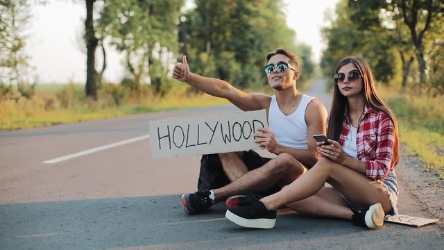 A young couple are hitchhiking sitting on the road and using smartphone. A man and a woman stop the car on the highway with a sign Hollywood