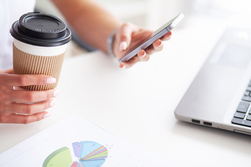 Woman using a smartphone and holding a cup of coffee in her office