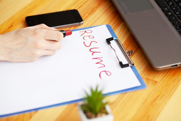 Job search. Female hand writes resume with lipstick on white sheet of paper. Wooden office desk with laptop, smartphone and supplies