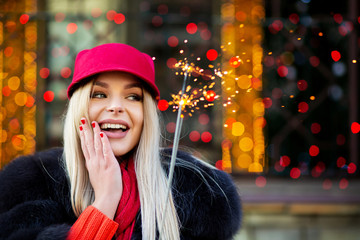 Classy blonde girl wearing fashionable winter outfit, having fun with sparklers at the bokeh background. Space for text