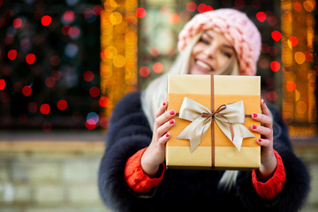 Charming blonde girl wearing fur coat and cap pulls a gift box to you. Space for text