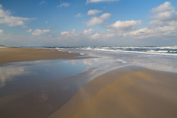 Vast beach at ebb tide, blue sky and white clouds reflected in the water of a gully
