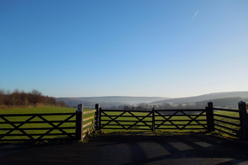 Fototapeta na wymiar Beautiful misty fields of the Derbyshire Dales on a sunny winter day, with a wooden fence in the foreground and a vapor trail in the sky.
