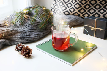 Cozy Christmas morning breakfast scene. Steaming glass cup of hot fruit tea standing near window on note pad. Glittering lights, pine cones, gift box and Christmas tree branch on wool plaid.