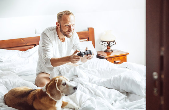 Adult man waked up and plays PC games sitting in bed, his dog watches on him with big interest