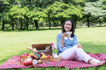 Elderly lifestyle concept. Attractive senior woman 50s holding white cup of coffee or tea have leisure time with picnic in the public park on the morning.
