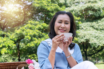 Fototapeta premium Elderly lifestyle concept. Attractive senior woman 50s holding white cup of coffee or tea have leisure time with picnic in the public park on the morning.