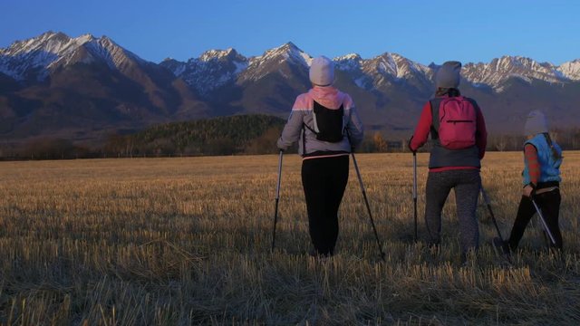 Woman Nordic walking in nature on background of mountains in field. Girls and children use trekking sticks and nordic poles, backpacks. Family travels sports. Kid is learning from mother and
