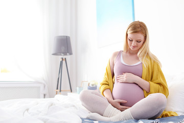 Beautiful pregnant woman sitting on bed in light room. Space for text