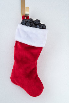 Traditional red and white plush Christmas stocking stuffed with coal shaped candy on a gold hook against a white background with silver flecks, as a naughty for Christmas concept background