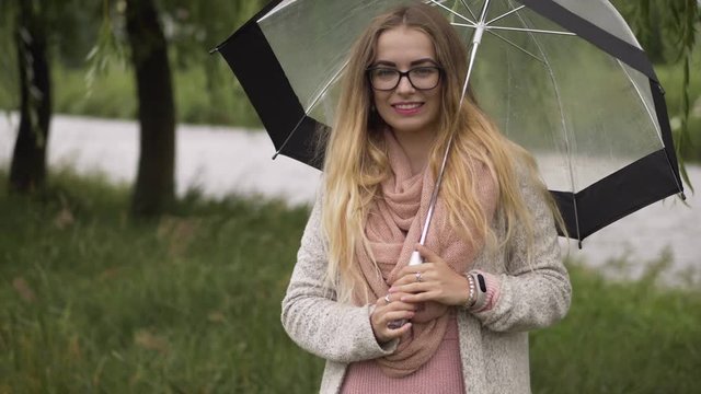Young beautiful blonde woman in cozy outfit with umbrella having fun strolling city park on a rainy and windy day