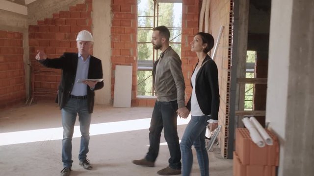 Sales agent talking with clients in new building. Engineer working in construction site with customers. Architect showing home to man and woman. Young couple as buyers of new house