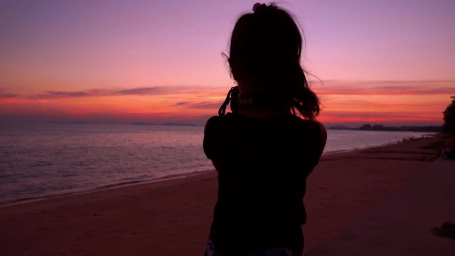 Silhouette little girl using camera taking photo on beach in sunset with twilight beautiful sky, slow-motion