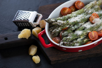 Baked asparagus with cherry tomatoes and grated parmesan on a black wooden serving board, studio shot