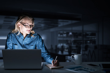 Attractive blonde working on laptop in dark office. Mixed media