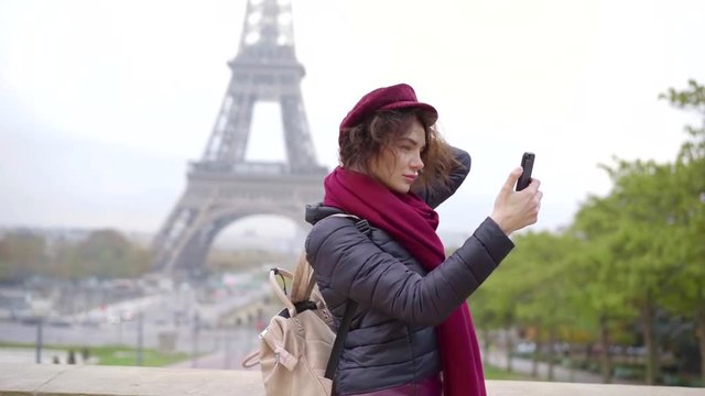 beautiful parisian girl is taking selfies against Eiffel tower in autumn day, correcting her cap