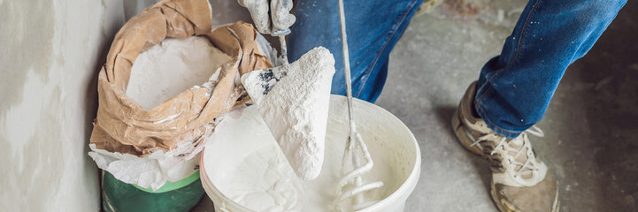 Young male painter kneads putty with water in a bucket using a hand-held mixer for building mixes...