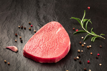 A photo of a steak of eye round beef, a raw cut, with rosemary, pepper and garlic on a black background with copy space