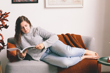 Young woman reading the magazine. Girl lying on the sofa. Woman holding the book in hands