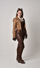 full length portrait of brunette  girl wearing brown leather steampunk outfit. standing pose, on grey studio background.