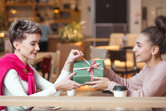 Happy caucasian woman congratulates her female friend with present box while sitting in mall cafe. Female friendship, girlfriends, holidays celebrating concept