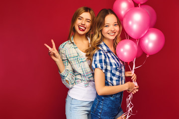 Two smiling beautiful women in hipster clothes .Girls posing on red background.Models with pink balloons.Having fun,ready for celebration birthday  or holiday party