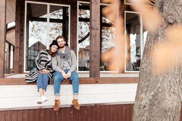 Young couple dressed in stylish warm coats sitting with cups on the terrace of the modern house...