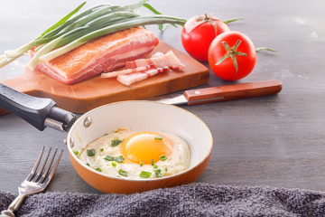 Fried egg in a pan, bacon, tomatoes and green onions are cooked for breakfast on a wooden gray table.