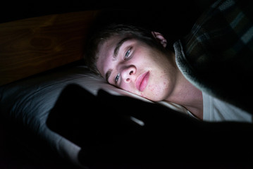 Depressed teenager browsing the internet on his mobile phone as he is lying on his bed in the dark.