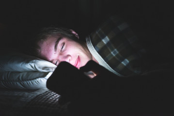 Teenager watching an exciting video on his mobile phone as he is lying on his bed in the dark.