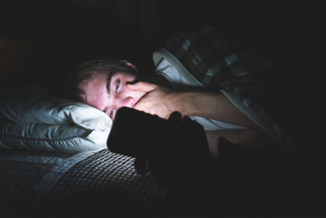Teenager reading some shocking news on his cell phone as he is lying on his bed in the dark.