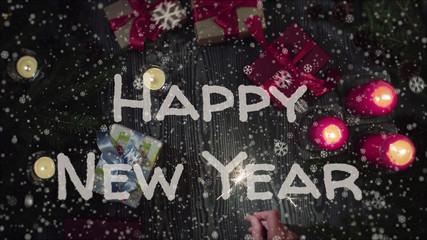 Greeting card Happy New Year, white letters, red candles and gifts, falling snow. Top view