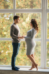 Happy family man and pregnant woman stand in front of big window in their house. They wait for a baby