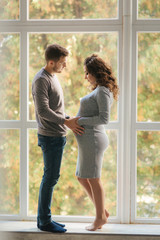Happy family man and pregnant woman stand in front of big window in their house. They wait for a baby