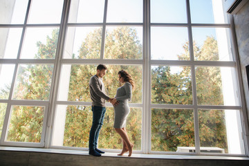 Beautiful couple stand in front of big window in studio. Pregnant woman with her husband