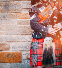 EDINBURGH, SCOTLAND, 24 March 2018 , Scottish bagpiper dressed in traditional red and black tartan dress stand before stone wall. Edinburgh, the most popular tourist city destination in Scotland. - obrazy, fototapety, plakaty