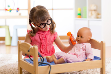 Child girl playing doctor examining baby doll patient with toy otoscope