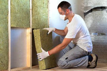 Worker in protective goggles and respirator insulating rock wool insulation in wooden frame for...