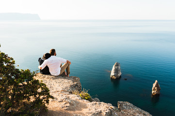 Loving couple sitting on the edge of the cliff by the sea. Honeymoon. Honeymoon trip. Boy and girl at the sea. Man and woman traveling. Couple hugs. Couple kissing. Newly married couple. Lovers