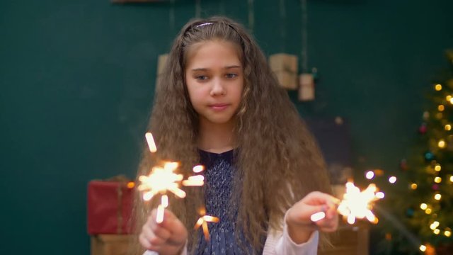 Cheerful happy litte girl looking with admiration at burning sparklers in her hands over Christmas background. Beautiful preadolescent child holiding burning bengal lights in christmas decorated room.