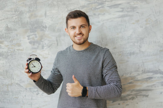Handsome man with beard stand on the gray background in studio. He hold a clock. Man show like