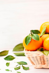 Fresh tangerines with green leaves in a wicker basket on a light wooden background.